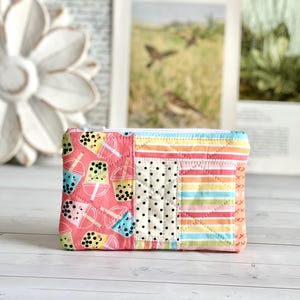 Peut inclure: Une pochette rectangulaire matelassée avec fermeture éclair. Le design patchwork présente des motifs variés, dont des tasses de thé boba sur fond rose, des pois et des rayures colorées. Idéal pour transporter de petits objets.