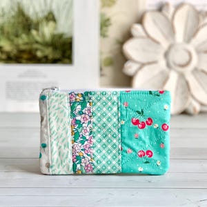 May include: A small, rectangular quilted pouch with a zipper closure. The pouch is made of fabric strips in various patterns, including floral, geometric, and cherry designs. The color palette includes white, light green, and pink.