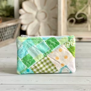 May include: A rectangular zippered pouch featuring a quilted patchwork design. The fabric panels include shades of green, light blue, and cream, with patterns such as gingham, polka dots, and linear textures, finished with a silver metal zipper.