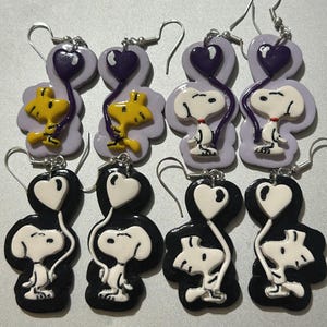 May include: A collection of handmade earrings featuring cartoon characters. The earrings showcase Snoopy and Woodstock designs, with heart-shaped balloons. The color palette includes black, white, yellow, purple, and silver.