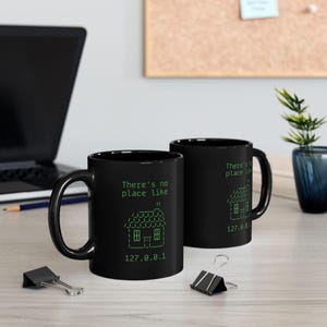 May include: Two black coffee mugs with a green text design. The text reads "There's no place like 127.0.0.1" and features a house icon.