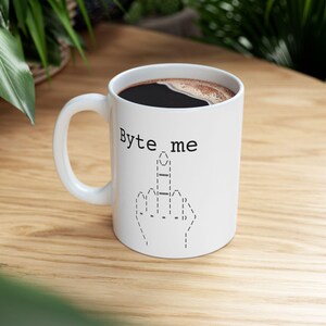 May include: White ceramic coffee mug with the text "Byte me" and a graphic of a hand giving the middle finger. The mug is filled with dark coffee and has a white handle. The mug is on a wooden surface.