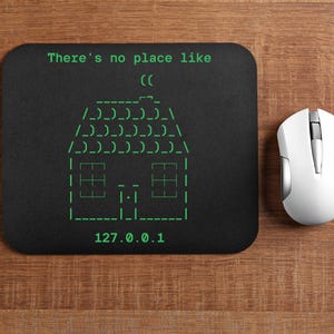 May include: Black rectangular mousepad with a green ASCII art house and the text "There's no place like" and "127.0.0.1". A white wireless mouse is next to the mousepad on a wooden surface.