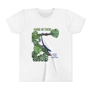 May include: A white short-sleeve t-shirt featuring a graphic of a blue tree monitor lizard hanging from a brown branch surrounded by green monstera leaves. The text HANG IN THERE is printed in green above, and BLUE TREE MONITOR in blue below.