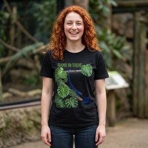 May include: A black short-sleeved t-shirt featuring a graphic of a blue tree monitor lizard clinging to a branch surrounded by green monstera leaves. The text HANG IN THERE is printed above the lizard, and BLUE TREE MONITOR is printed below.