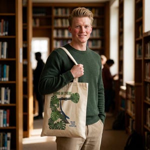 May include: A beige canvas tote bag featuring a graphic print of green monstera leaves and a blue lizard on a branch. The text HANG IN THERE and BLUE TREE MONITOR is printed in dark green and blue ink on the front of the bag.