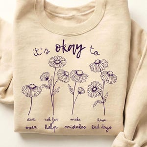 Könnte beinhalten: Sandfarbenes Sweatshirt mit dem Text "it's okay to" über einem Blumendesign. Unter den Blumen stehen die Worte "start over, ask for help, make mistakes, have bad days". Das Wort "SAND" befindet sich in der oberen rechten Ecke.