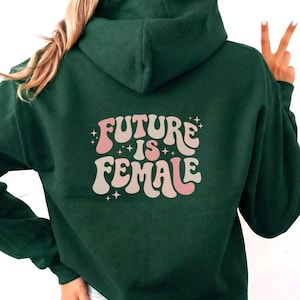 Może przedstawiać: Ciemnozielona bluza z kapturem z napisem "FUTURE IS FEMALE" w stylu retro na plecach. Tekst jest w kolorze różowym i białym. Bluza ma kaptur i przednią kieszeń.