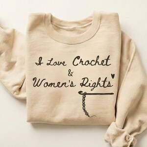 Könnte beinhalten: Sandfarbenes Sweatshirt mit dem Text "I Love Crochet & Women's Rights" in schwarzer Schrift. Das Design beinhaltet einen Häkelhaken und eine Kette. Die Ärmel sind hochgekrempelt. Das Wort "SAND" befindet sich in der oberen rechten Ecke.
