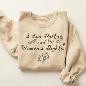 Puede incluir: Sudadera color arena con el texto negro "I Love Pasta and Women's Rights". El diseño incluye ilustraciones de formas de pasta. Las mangas están enrolladas. La palabra "SAND" está en la esquina superior derecha.