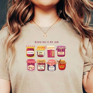 Könnte beinhalten: Beigefarbenes T-Shirt mit dem Text "BOOKTOK IS MY JAM" und einer Grafik von neun Marmeladengläsern. Jedes Glas hat ein anderes Etikett, darunter "Cozy Reads", "Slow Burn" und "Dark Romance".