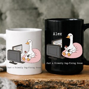 May include: Two ceramic mugs, one white and one black, with a humorous design. Each mug features a cartoon goose sitting on a pink cushion in front of a computer. The text "Just a Friendly Bug-Fixing Goose" is printed on both mugs. The black mug has the name "Alex" printed above the goose.