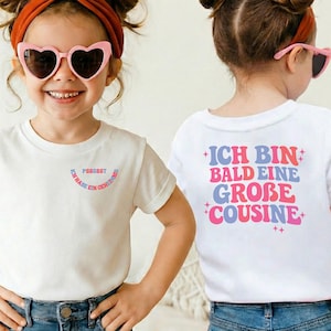 Puede incluir: Una camiseta blanca con un pequeño gráfico frontal con el texto PSSSSST ICH HABE EIN GEHEIMNIS y un gráfico grande en la espalda con el texto ICH BIN BALD EINE GROBE COUSINE en letras onduladas rosas y azules con pequeños detalles de estrellas.