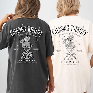 Könnte beinhalten: Zwei kurzärmelige T-Shirts, eines in Anthrazit und eines in Creme, jeweils mit einer schwarzen Grafik auf der Rückseite. Das Design zeigt ein Skelett in einem Martiniglas mit dem Text "CHASING TOTALITY," "Summer of Darkness," "SPAIN 2026," "AUGUST 12TH," und "TOTAL SOLAR ECLIPSE" sowie Mondphasen.