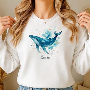 Könnte beinhalten: Weißes Sweatshirt mit einer Aquarellillustration eines Blauwals. Der Wal ist in Blautönen vor einem türkisfarbenen Hintergrund dargestellt. Der Name "Laura" ist darunter geschrieben. Ein lässiger, bequemer Stil.
