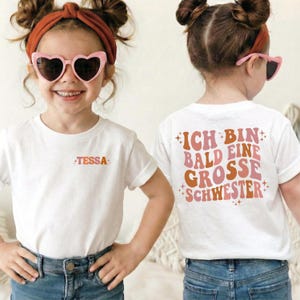 Puede incluir: Una camiseta blanca de manga corta con el nombre Tessa en naranja en el pecho izquierdo. La parte trasera muestra el texto Ich bin bald eine grosse Schwester en letras onduladas de color rosa y naranja con estrellas.