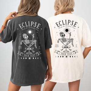 Könnte beinhalten: Ein anthrazitfarbenes T-Shirt und ein cremefarbenes T-Shirt, beide mit einer Grafik auf der Rückseite, die ein Skelett in einem Martiniglas zeigt, mit dem Text ECLIPSE, Make the Darkness Count, SOLAR ECLIPSE 20 26 und YOUR TEXT auf einem Banner.