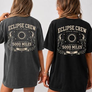 May include: A charcoal gray t-shirt featuring a back graphic with the text ECLIPSE CREW SPAIN 2026, I TRAVELED 5000 MILES FOR 90 SECONDS, and a name banner. The design includes a central solar eclipse, crescent moons with ribbons, and stars.