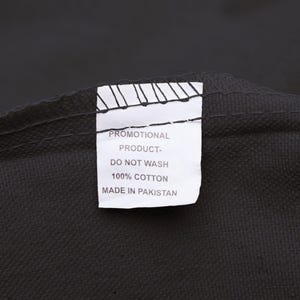 May include: Close-up of a black textile with a white tag. The tag reads "PROMOTIONAL PRODUCT - DO NOT WASH - 100% COTTON - MADE IN PAKISTAN". The fabric appears to be a dark, solid color.