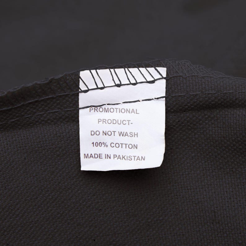 May include: Close-up of a black textile with a white tag. The tag reads "PROMOTIONAL PRODUCT - DO NOT WASH - 100% COTTON - MADE IN PAKISTAN". The fabric appears to be a dark, solid color.