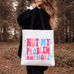 Może przedstawiać: Biała torba typu tote z czarnymi paskami na ramię, z napisem NOT MY PROBLEM ANYMORE wykonanym falowaną, wielokolorową czcionką w odcieniach różu, jasnego różu i jasnego błękitu, ozdobioną małymi czteroramiennymi gwiazdkami w kolorze niebieskim i różowym.