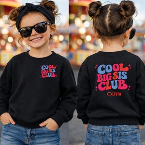 May include: Black sweatshirt featuring the text "COOL BIG SIS CLUB" in blue, red, and pink. The sweatshirt is shown from the front and back. The back of the sweatshirt also has the name "OLIVIA" printed below the text.
