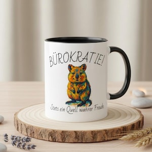 Könnte beinhalten: Weißer Keramikbecher mit schwarzem Innenraum und Henkel. Der Becher zeigt eine farbenfrohe Illustration eines Quokkas und den deutschen Text "Bürokratie!" über dem Quokka, mit dem Text "Stets ein Quell wahrer Freude" darunter.