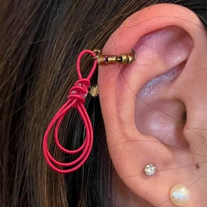 May include: A close-up of an ear adorned with a vibrant pink ear cuff and a gold-toned ear wrap. The ear also features a small diamond stud and a pearl earring. The pink ear cuff has a looped design.