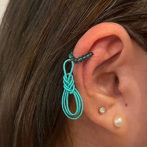 May include: A turquoise ear cuff with a woven design and a beaded upper section. The ear cuff is worn on the ear, with a small stud earring and a pearl earring also visible.