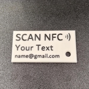 Op de afbeelding: Een witte rechthoekige NFC-tag met zwarte tekst met de tekst "SCAN NFC ))", "Your Text" en "name@gmail.com". Er zit een klein gaatje in de hoek. De tag ligt op een donkergrijze ondergrond.