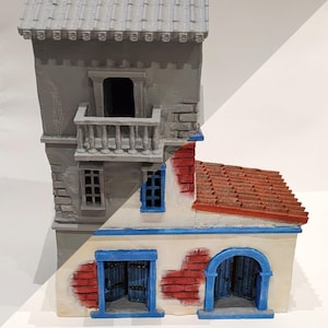 Op de afbeelding: Een model van een gebouw met twee verdiepingen, geschilderd in een realistische stijl. De begane grond is wit met rode bakstenen accenten en blauwe deuren. De bovenverdieping is grijs met een balkon en ramen. Het dak is van rode tegels.