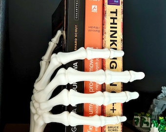 May include: A white skeleton hand bookend holding several books. The bookend is made of a hard material and has a rectangular base. The books have various colors and titles, including "THINKING" and "GALAKTYKA".