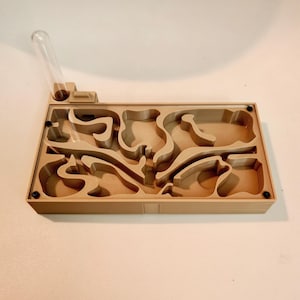 May include: A tan ant farm with a complex internal structure, featuring winding pathways and chambers. A clear glass tube is connected to the side. The rectangular design includes black screws at the corners.