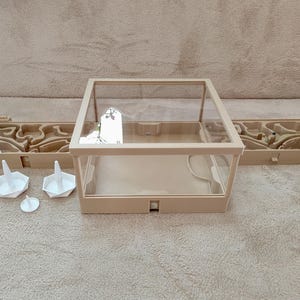 May include: A beige ant farm with a clear acrylic viewing box and two side chambers with a maze-like design. Includes white feeding dishes and a small, round water source. The ant farm is designed for observing ants.