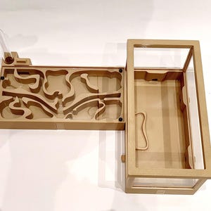 Könnte beinhalten: Eine beige Ameisenfarm mit einem transparenten Kunststoffrohr und einer rechteckigen Kammer mit labyrinthartigem Design. Die Farm hat eine separate, größere Kammer mit einer transparenten Glasplatte. Das Design ist modern und funktional.