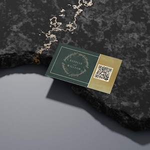 May include: A business card with a dark green background and gold accents. The card features the names "Estelle" and "William" inside a wreath design. A QR code is on the right side of the card. The card is placed on a dark, textured surface.