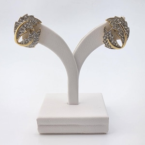May include: A pair of leaf-shaped earrings. Each earring features a gold-tone frame with a leaf design encrusted with small, clear crystals. The earrings are displayed on a white jewelry stand against a white background.
