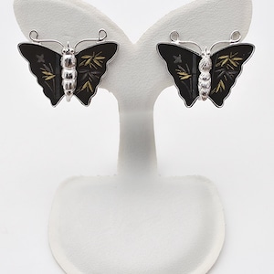 May include: A pair of butterfly earrings. Each earring features a black enamel butterfly with silver-colored metal accents. The butterfly wings have gold and gray leaf-like details. The earrings are displayed on a white stand.