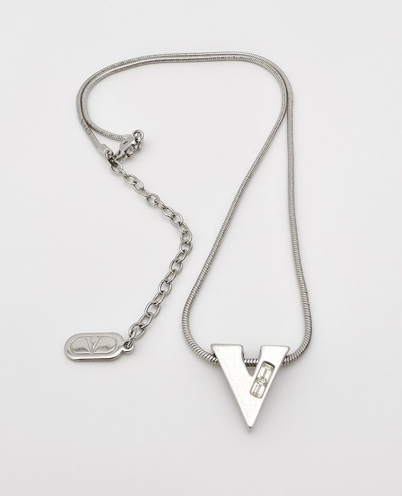 May include: A silver chain necklace with a silver pendant shaped like the letter V. The pendant has a small clear crystal in the center.