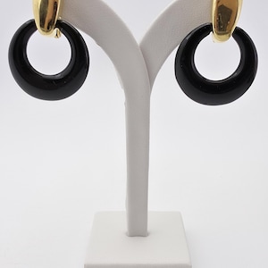 以下が含まれることがあります： ゴールドカラーのアクセントが付いた、黒いフープイヤリングのペア。イヤリングは白い背景に白いジュエリースタンドに展示されています。フープは光沢のある黒色です。