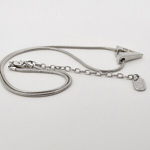 May include: A silver chain necklace with a silver pendant shaped like a triangle. The pendant has a small, round charm attached to it.
