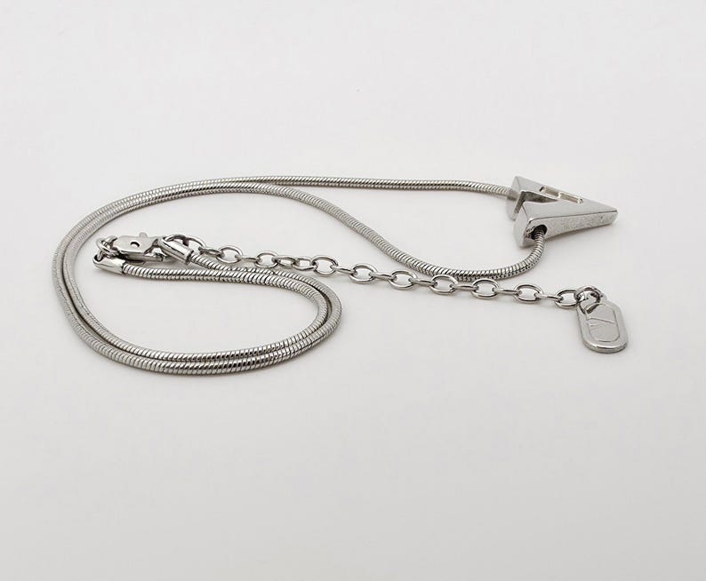 May include: A silver chain necklace with a silver pendant shaped like a triangle. The pendant has a small, round charm attached to it.