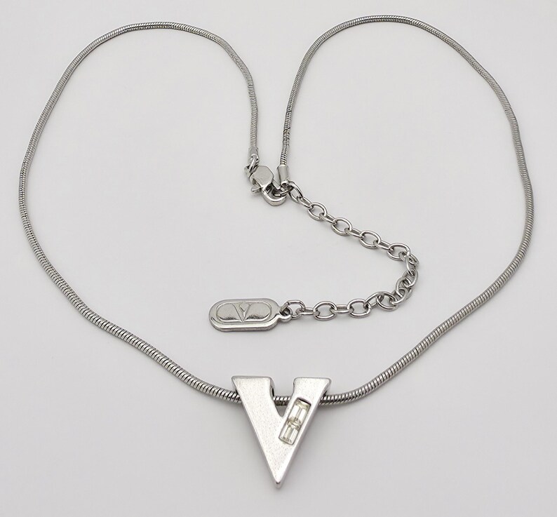 May include: A silver chain necklace with a silver pendant in the shape of the letter V. The pendant has a small clear stone in the center.