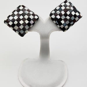 Pode incluir: Um par de brincos quadrados com um padrão xadrez em preto, branco e cinza. Os brincos são exibidos em um suporte branco sobre um fundo branco. Os brincos têm um acabamento brilhante.