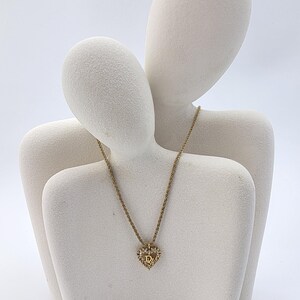 May include: A gold-toned necklace with a heart-shaped pendant. The pendant is encrusted with small, clear gemstones and features the letter "D" in the center. The necklace is displayed on a white, abstract sculpture.