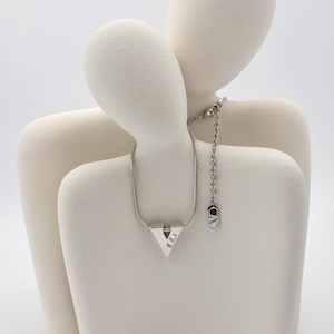 May include: A silver necklace with a V-shaped pendant featuring two small clear stones. The necklace is displayed on a white ceramic sculpture of two figures.