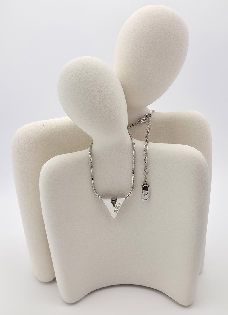 May include: A silver necklace with a V-shaped pendant featuring two small clear stones. The necklace is displayed on a white ceramic sculpture of two figures.
