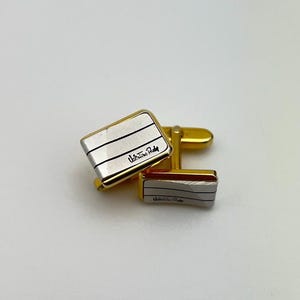 May include: A pair of gold cufflinks with a silver rectangular face. The face has three horizontal lines and the text "Valentino Rudy" in black script.