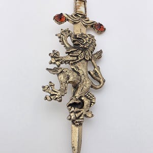 May include: A vintage gold-toned brooch featuring a lion rampant design on a sword. The sword's hilt has three red gemstone accents. The brooch is likely made of metal and is a decorative accessory.