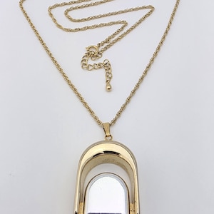 May include: A gold-toned necklace with a long chain and a pendant. The pendant features a gold arch design with a silver-colored rectangular insert. The necklace has an adjustable chain length.